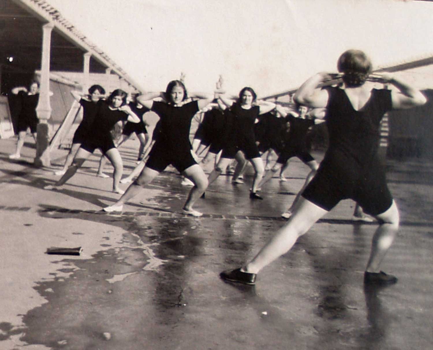Foto, schwarz-weiß, außen: Eine Gruppe von Menschen in schwarzen Turnanzügen macht einen Ausfallschritt zur Seite, das andere Knie ist gebeugt.