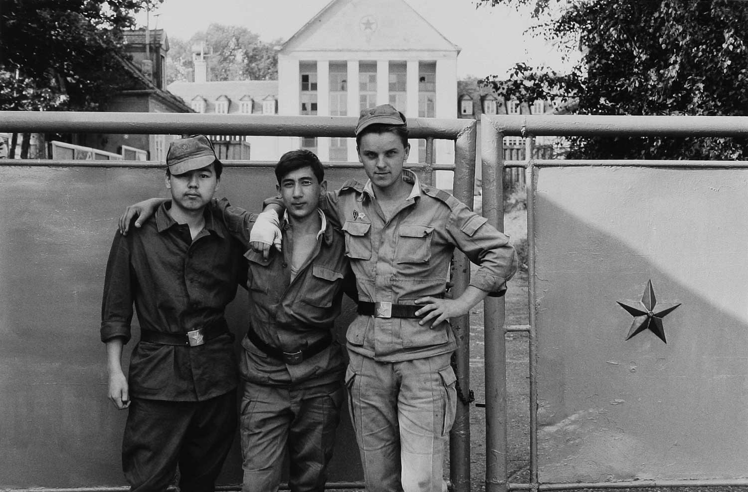 Foto, schwarz-weiß: Drei Soldaten in unterschiedlichen Uniformen vor dem geschlossenen Kasernentor aus Metall.