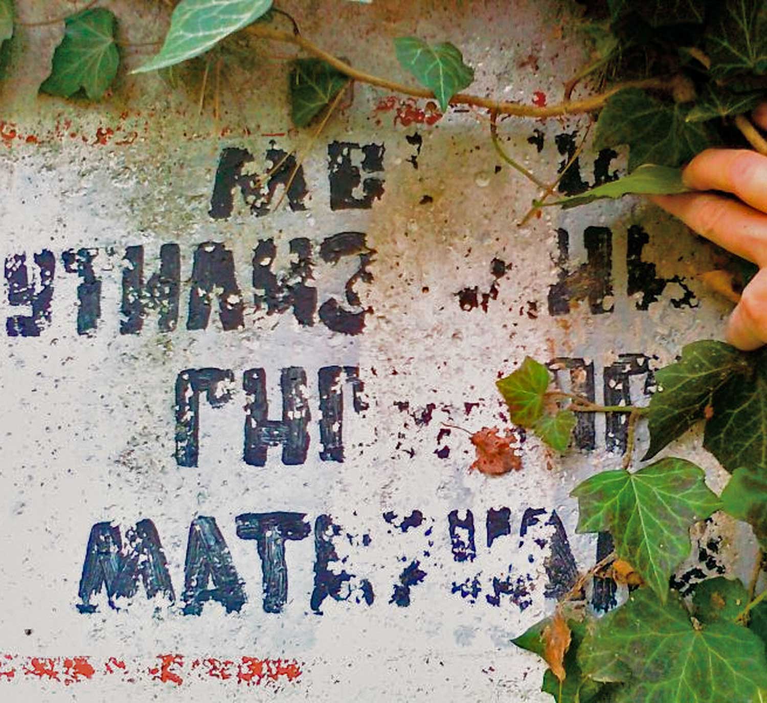 Foto, farbig: Nahaufnahme eines Mauerstücks mit aufgemalten, verwitterten kyrillischen Buchstaben hinter einer Efeuranke.