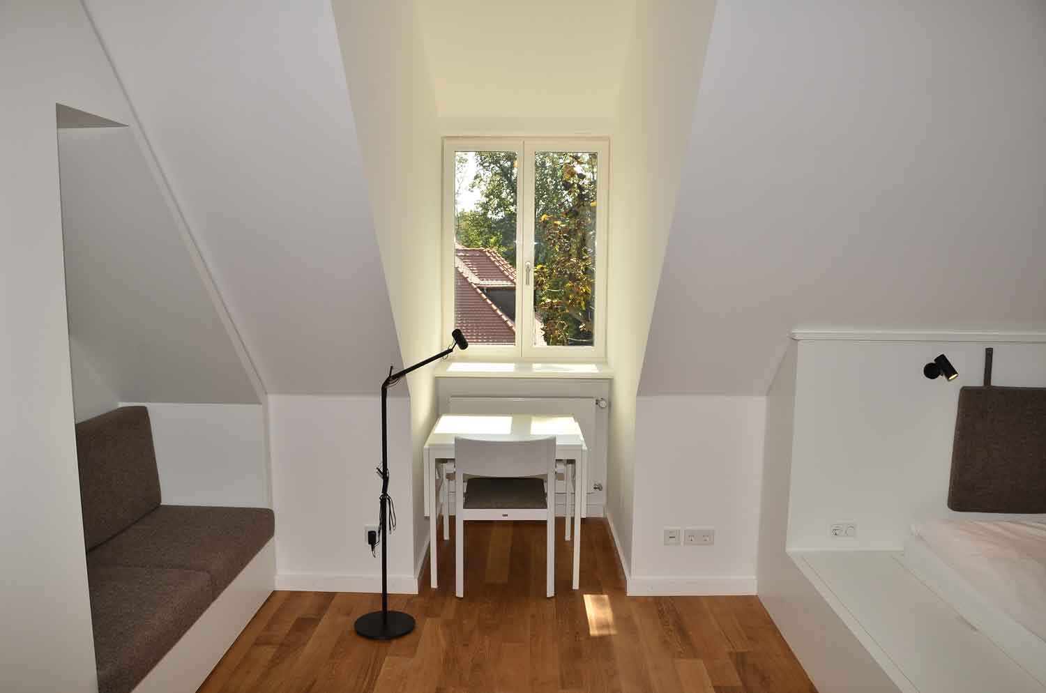 Foto, farbig, innen: Raum im Dachgeschoss mit weißen Wänden und Parkettfußboden, links eine Sitzbank mit schwarzem Bezug, in der Mitte eine Dachgaube mit kleinem Tisch und Stuhl, rechts eine Schlafgelegenheit.
