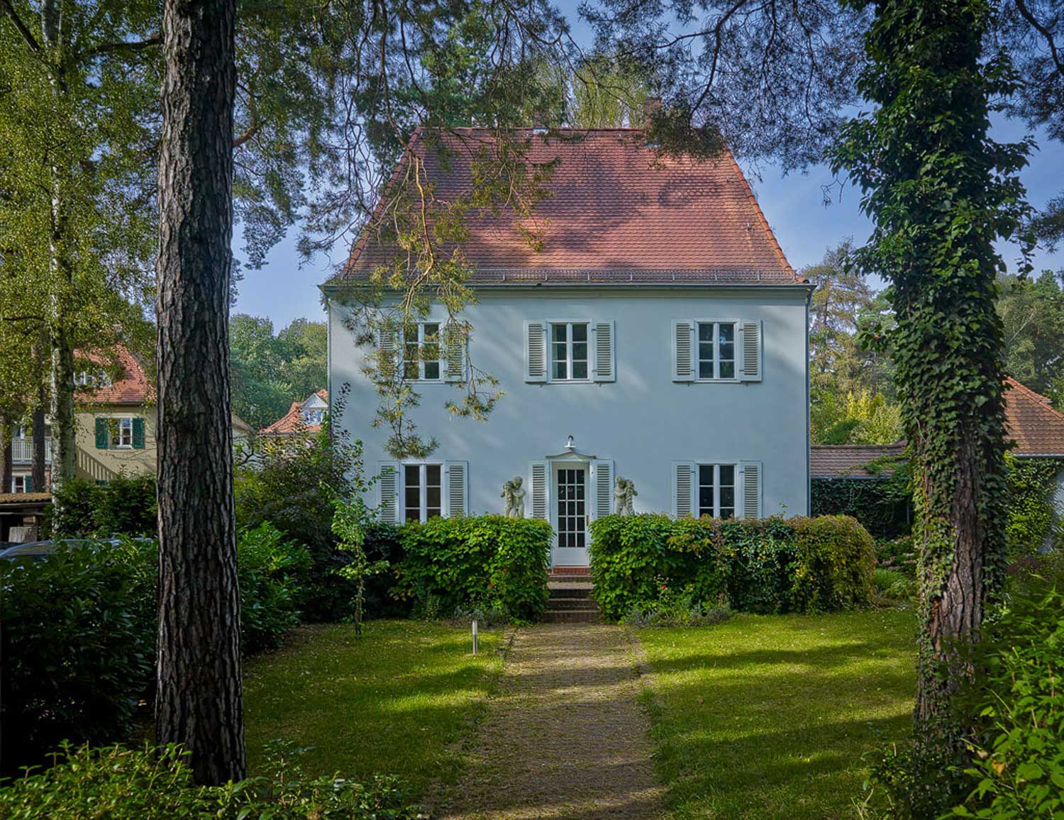 Foto, farbig, außen: Ein weißgestrichenes Haus mit Trapezdach, auf das ein Gartenweg hinführt, links und rechts ein belaubter Baum.