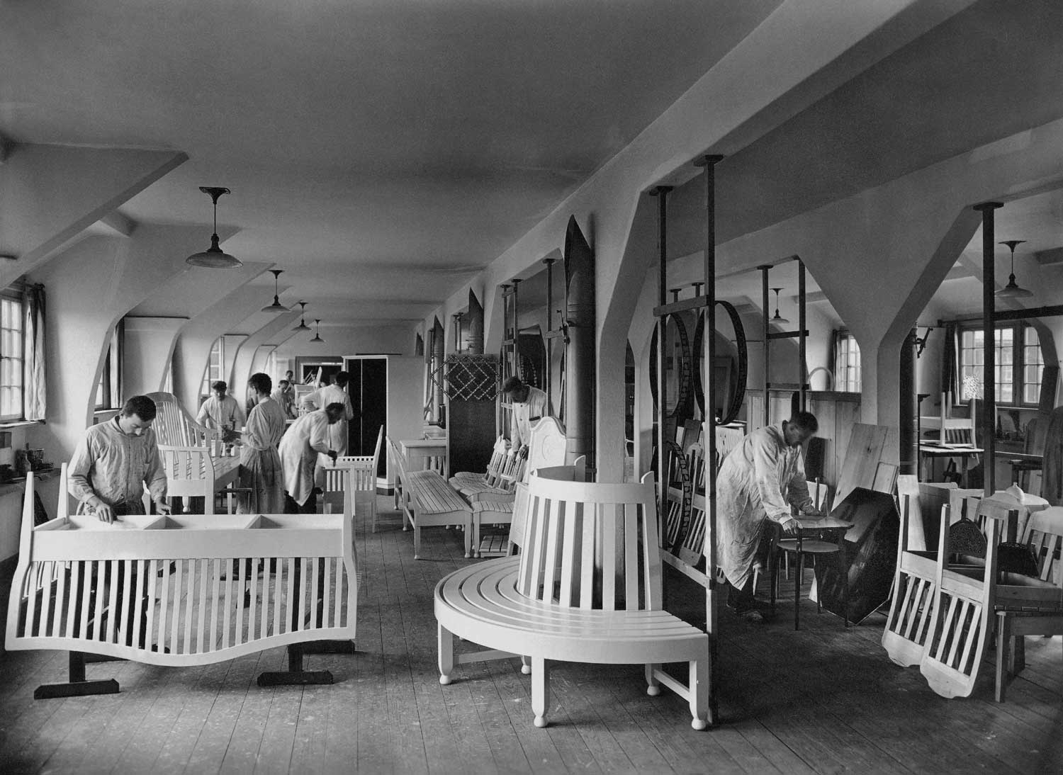 Foto, schwarz-weiß: Blick in eine Werkstatt, in der mehrere Menschen an Sitzmöbeln arbeiten.