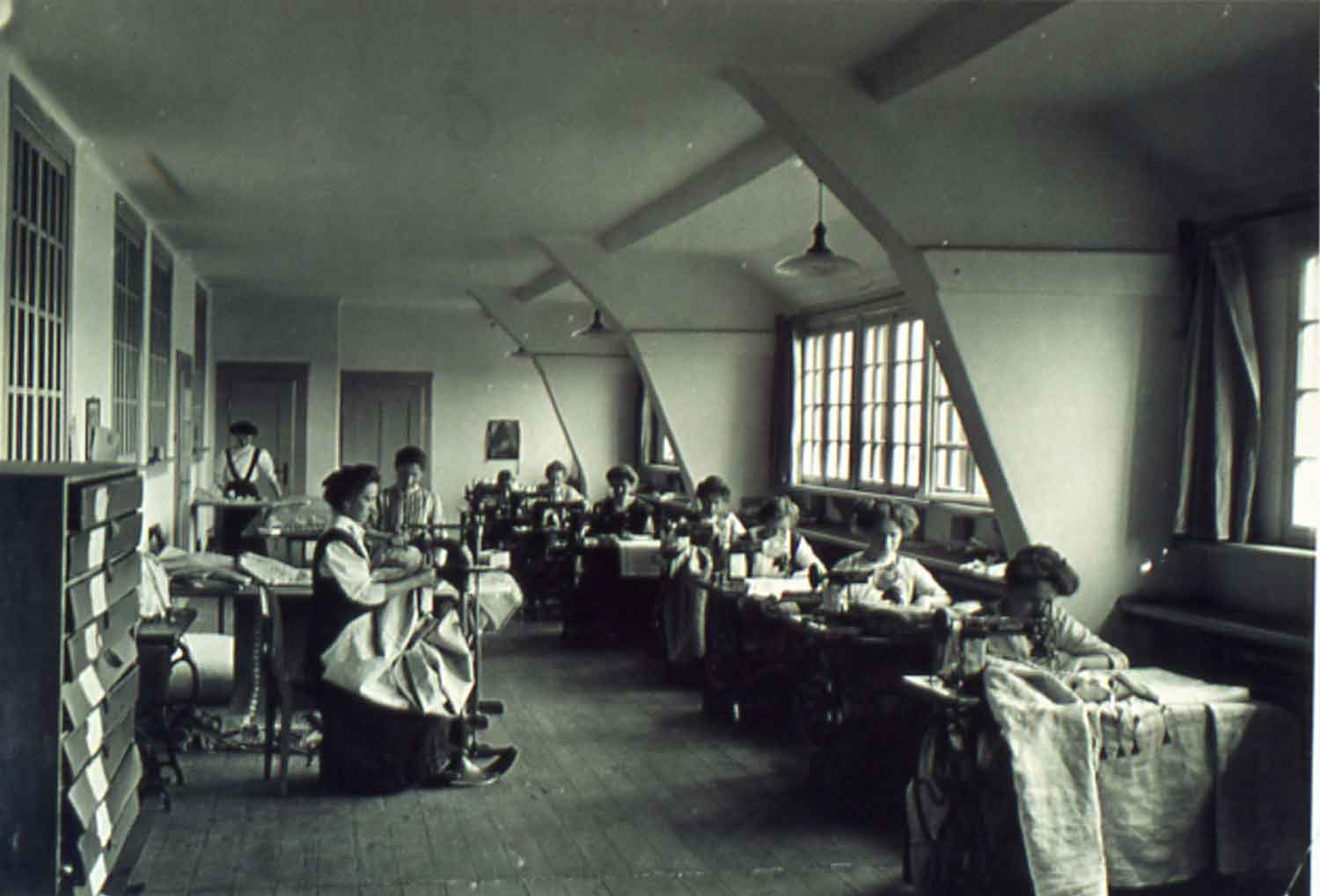 Foto, schwarz-weiß: In einem langgezogenen Raum sitzen rechts acht Frauen und links zwei Frauen hintereinander an Einzeltischen mit Nähmaschinen.