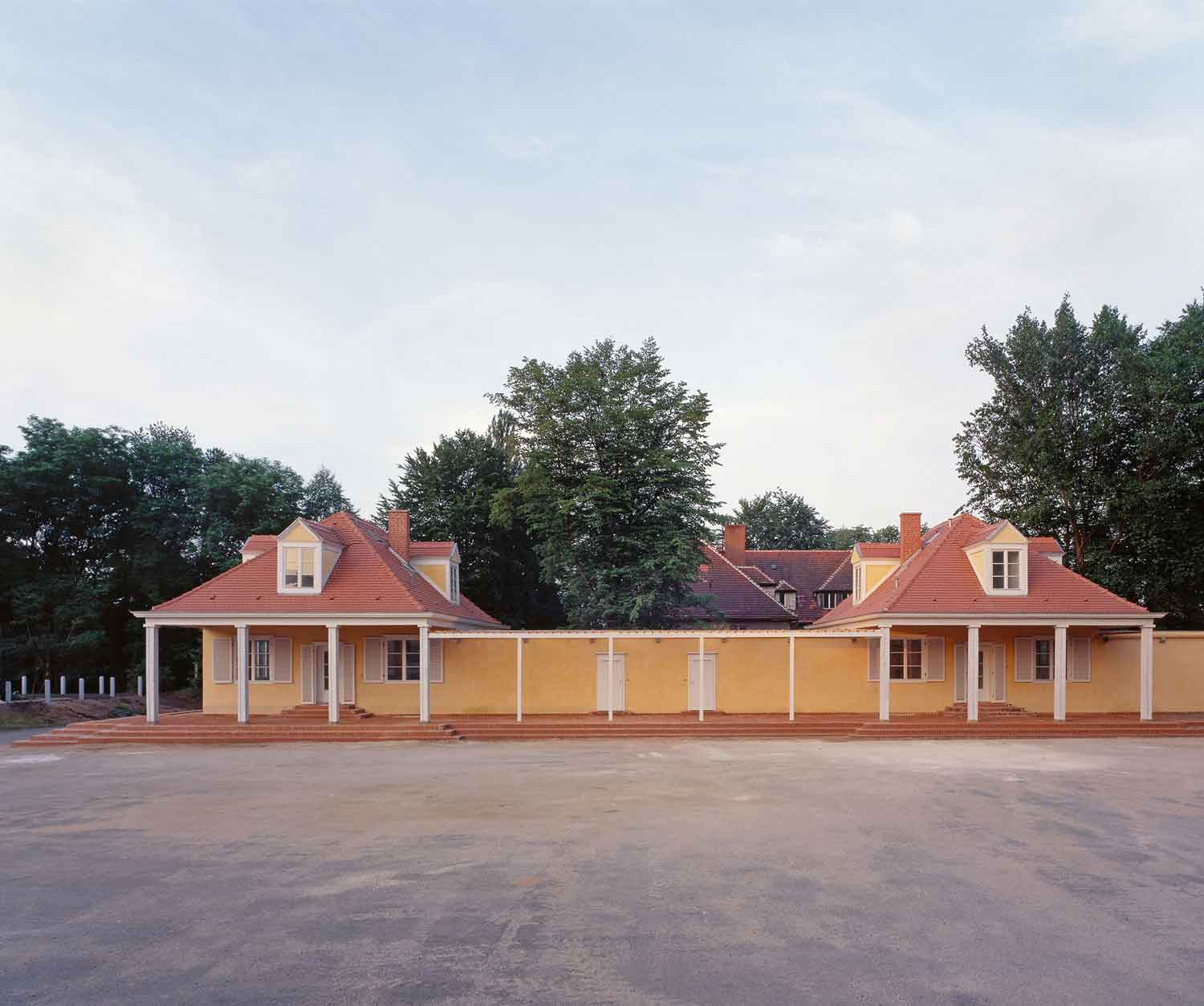 Foto, farbig: Frontalansicht der beiden kleinen, eingeschossigen Gebäude mit pyramidenförmigen Dächern, die durch einen Laubengang verbunden und gelb gestrichen sind.